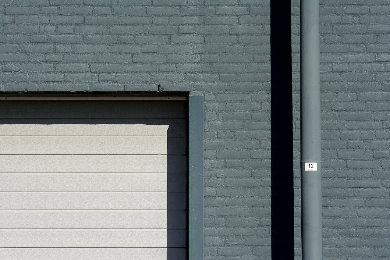 A minimalist image of a gray brick wall featuring a garage door and a numbered pipe.